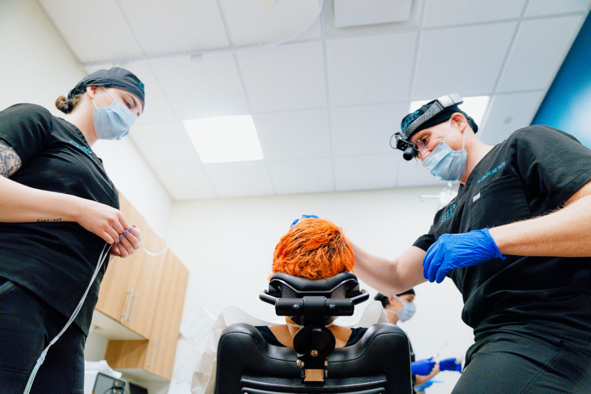 Two CMF professionals in black scrubs and magnification headgear attending to a seated patient with orange hair in a brightly lit clinical room.
