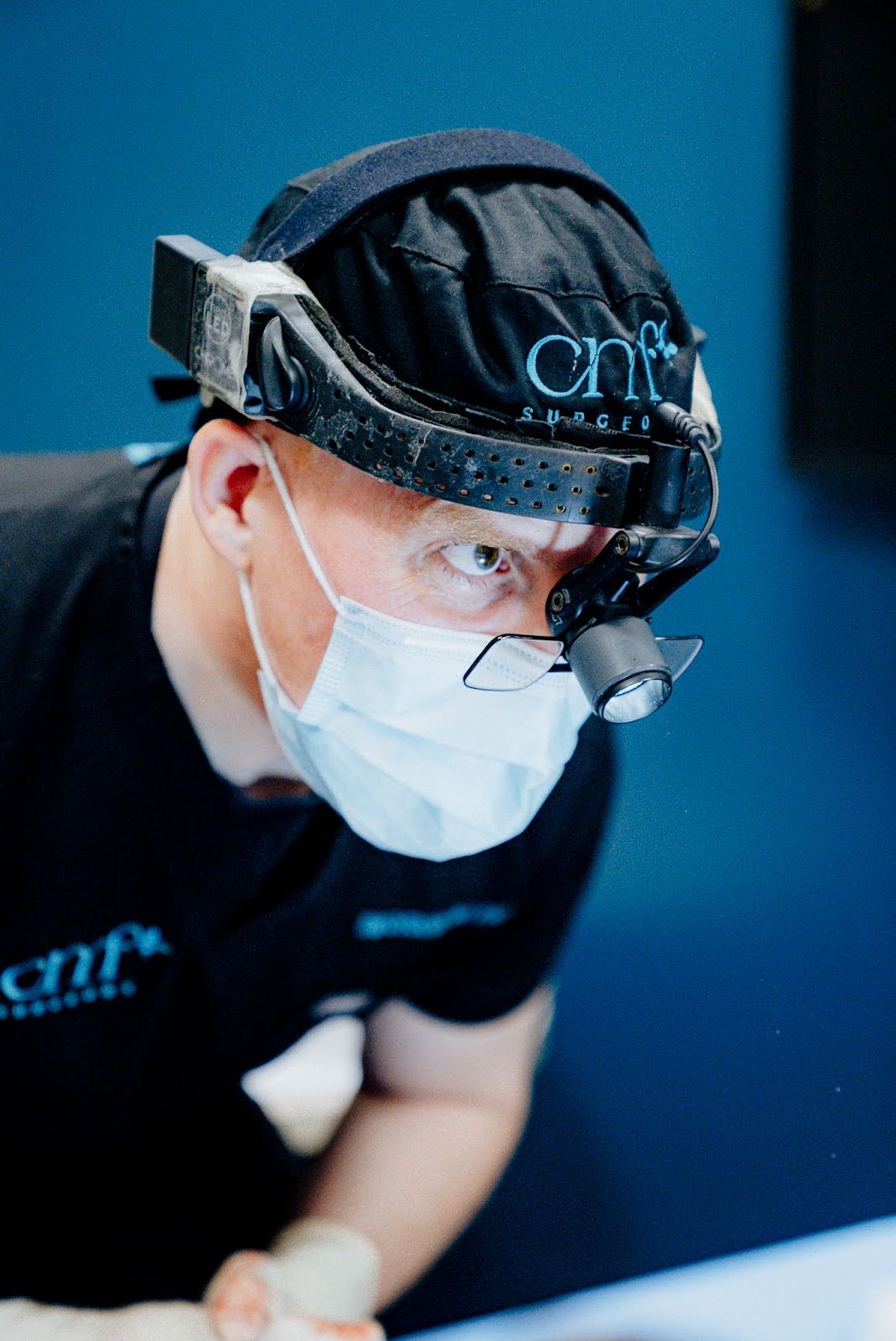 CMF surgeon wearing magnification headset and surgical mask, focused during a facial procedure, dressed in black scrubs with "CMF SURGEON" logo.