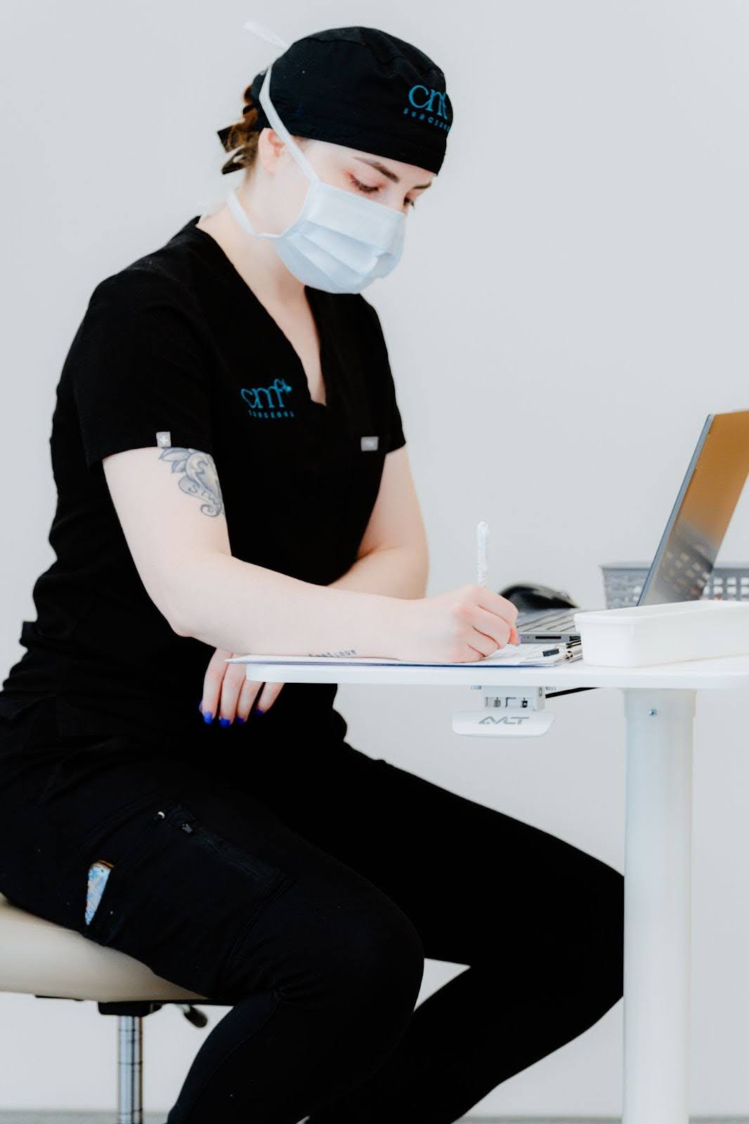 CMF medical professional in black scrubs and branded surgical cap, writing in a notebook while working at a laptop in a clinical office setting.